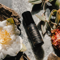 Photographers style a bottle of Oud Revele luxury essential oil blend among white flowers, leaves, and wood on a gray marble surface.
