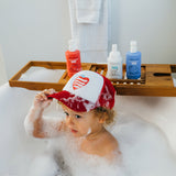 A toddler wearing a patriotic baseball cap plays in a bubbly bathtub filled with strawberry, vanilla, and blueberry bath products.
