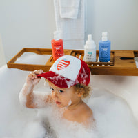A toddler wearing a patriotic baseball cap plays in a bubbly bathtub filled with strawberry, vanilla, and blueberry bath products.
