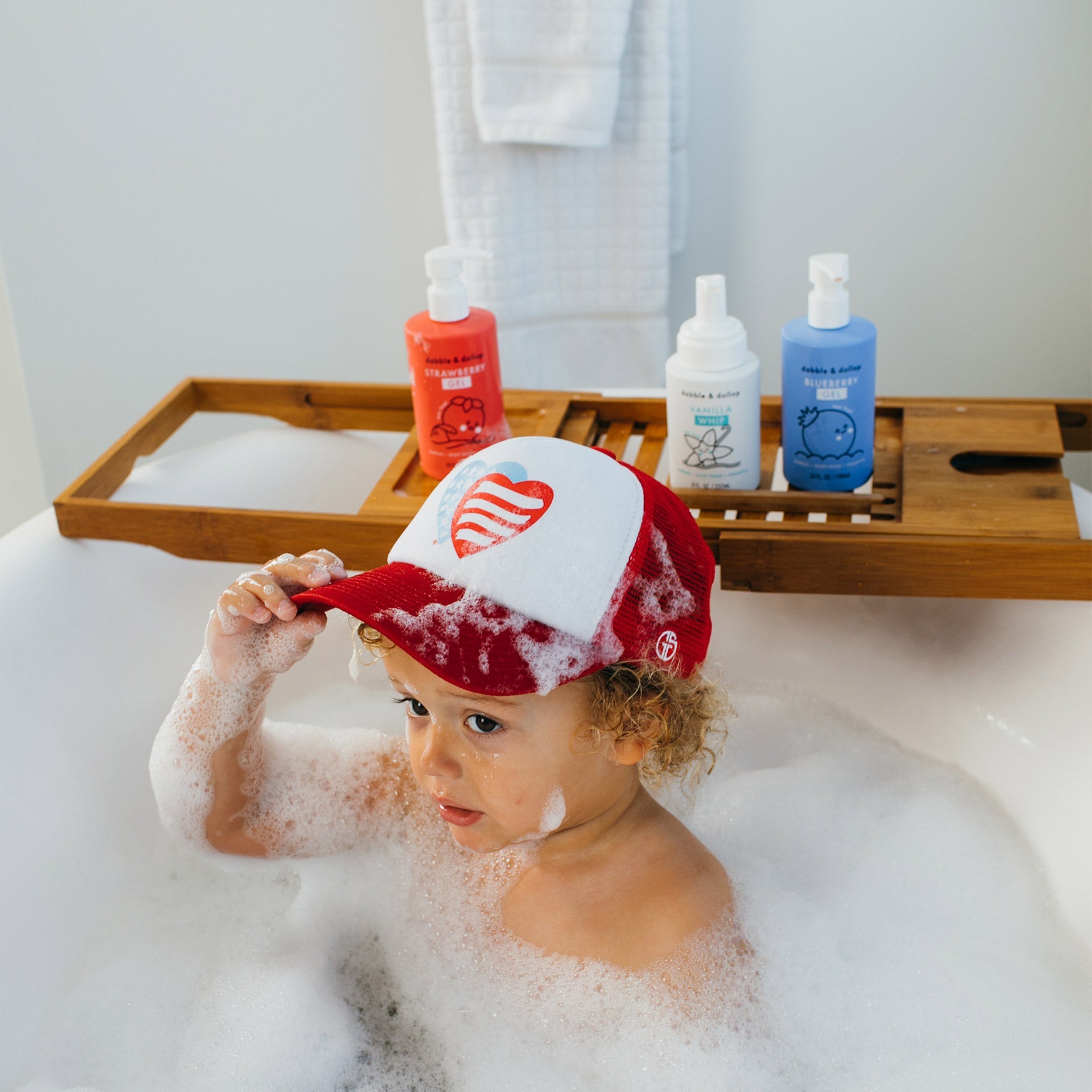 A toddler wearing a patriotic baseball cap plays in a bubbly bathtub filled with strawberry, vanilla, and blueberry bath products.
