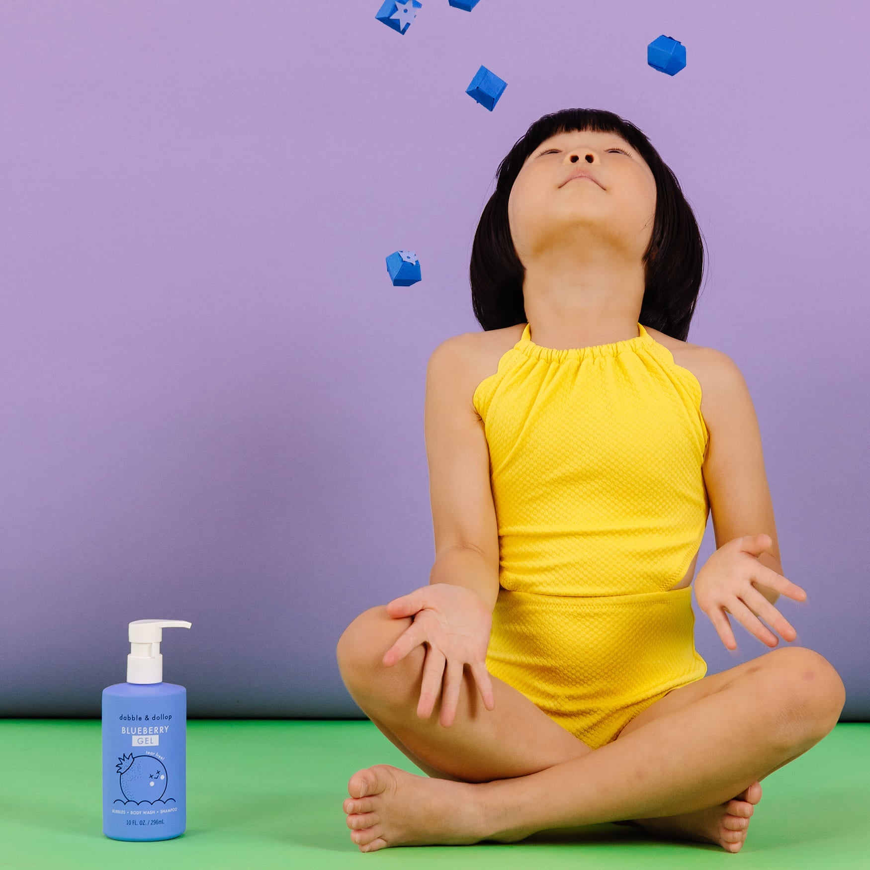 A young girl in a yellow swimsuit sits cross-legged and watches blue toy blocks fall near Dabble & Dollop Blueberry Gel body wash.
