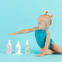 A young girl in a teal swimsuit reaches for Dabble & Dollop baby bath and shampoo products against a bright blue background.

