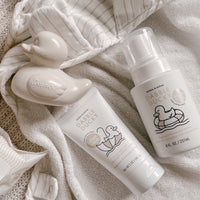 A baby bath scene displays Dabble Ducky baby wash, infant balm, and a rubber ducky on soft white towels.
