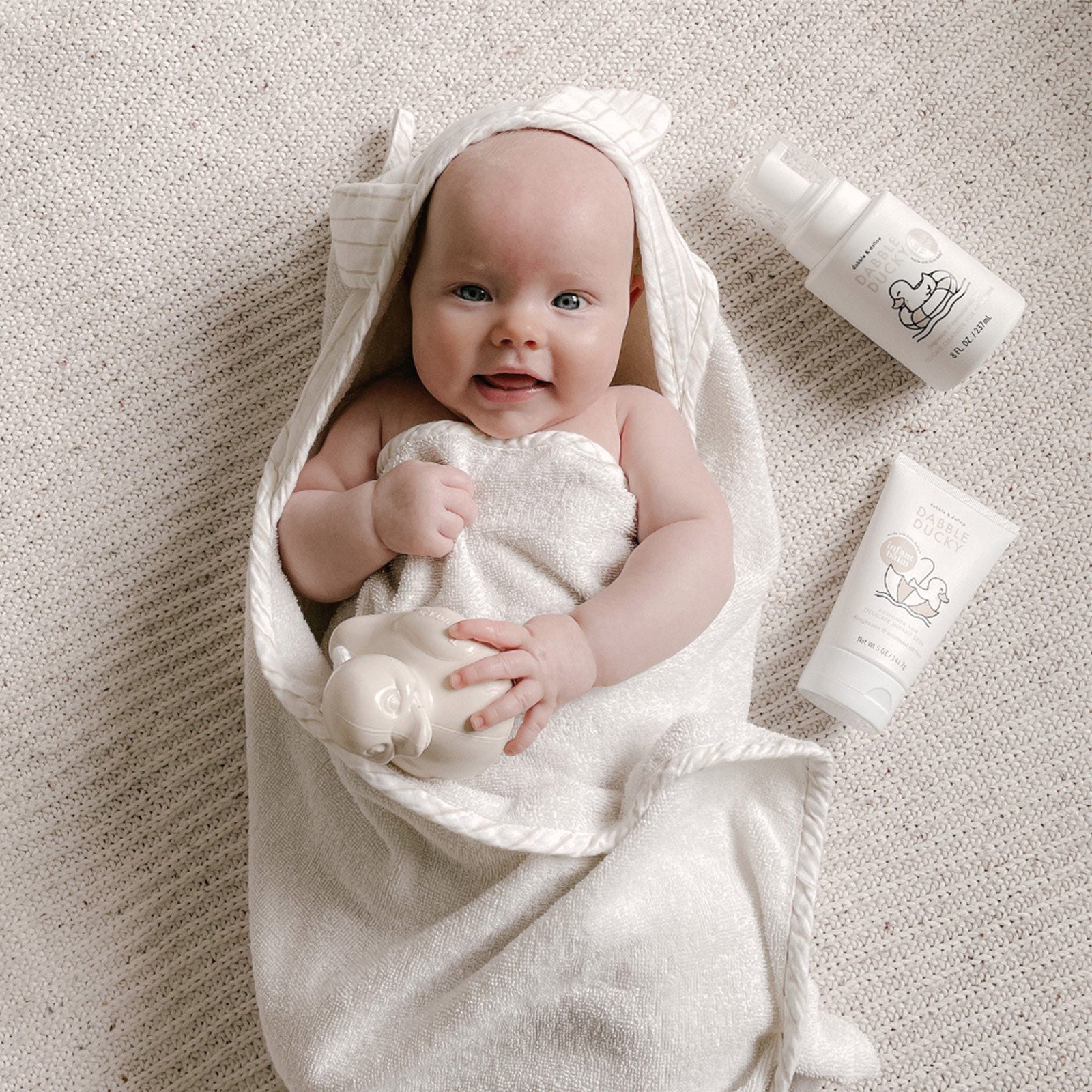 A smiling baby wrapped in a hooded towel holds a duck toy next to Dabble Ducky baby bath products.
