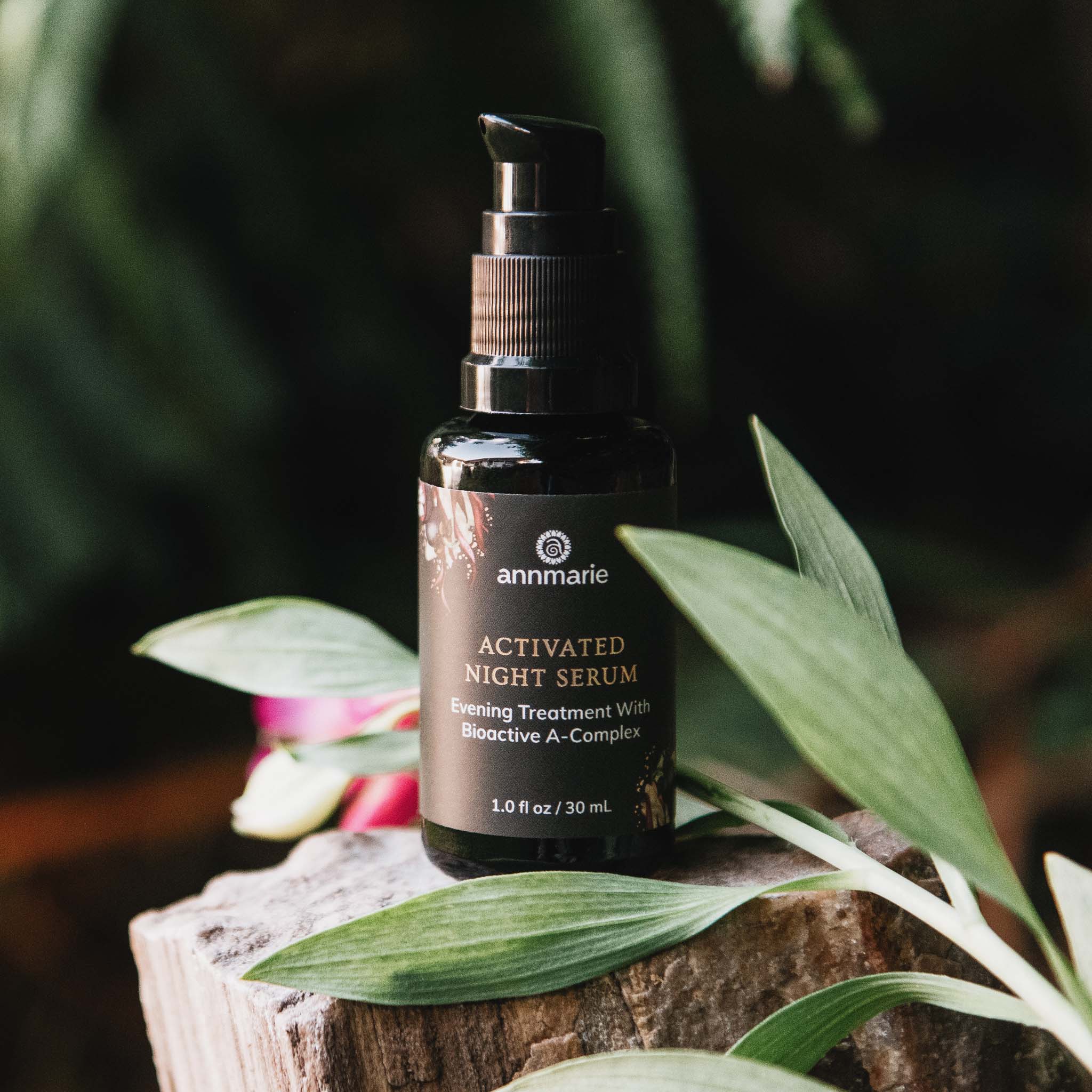 Annmarie Activated Night Serum sits atop a weathered wood block surrounded by green foliage, promoting natural skincare.
