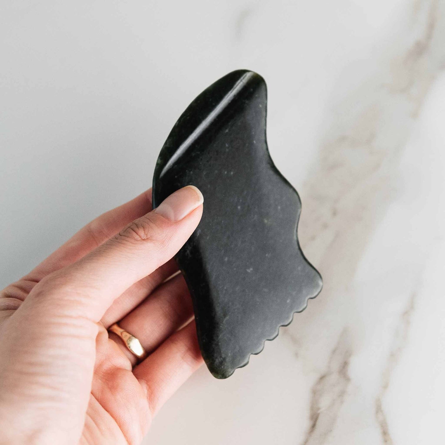Hand holds a dark green jade gua sha tool against a marble background.

