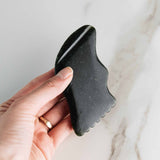 Hand holds a dark green jade gua sha tool against a marble background.
