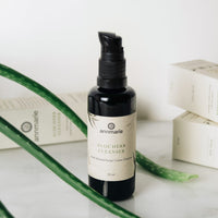 Annmarie's Aloe Herb Cleanser sits near fresh aloe vera stalks and product boxes showcasing the natural skincare line.
