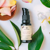 Annmarie Anti-Aging Facial Oil sits surrounded by leaves, rose petals, and a white iris flower.
