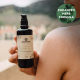A person displays a bottle of Annmarie Coconut Body Oil, highlighting its enhanced herb formula for natural skincare.
