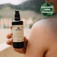 A person displays a bottle of Annmarie Coconut Body Oil, highlighting its enhanced herb formula for natural skincare.
