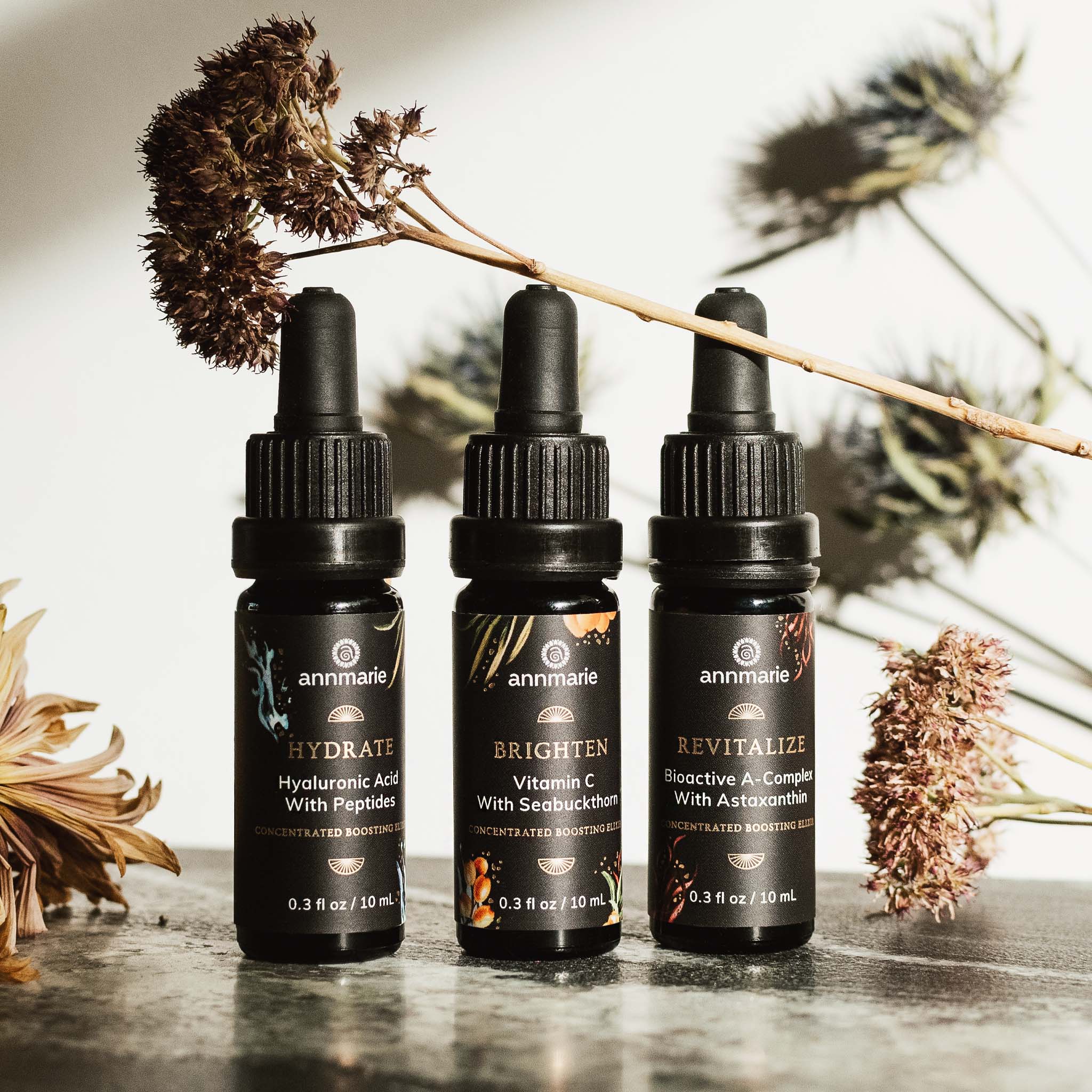 Annmarie's skincare collection presents three boosting elixirs – Hydrate, Brighten, and Revitalize – displayed against a backdrop of dried flowers.
