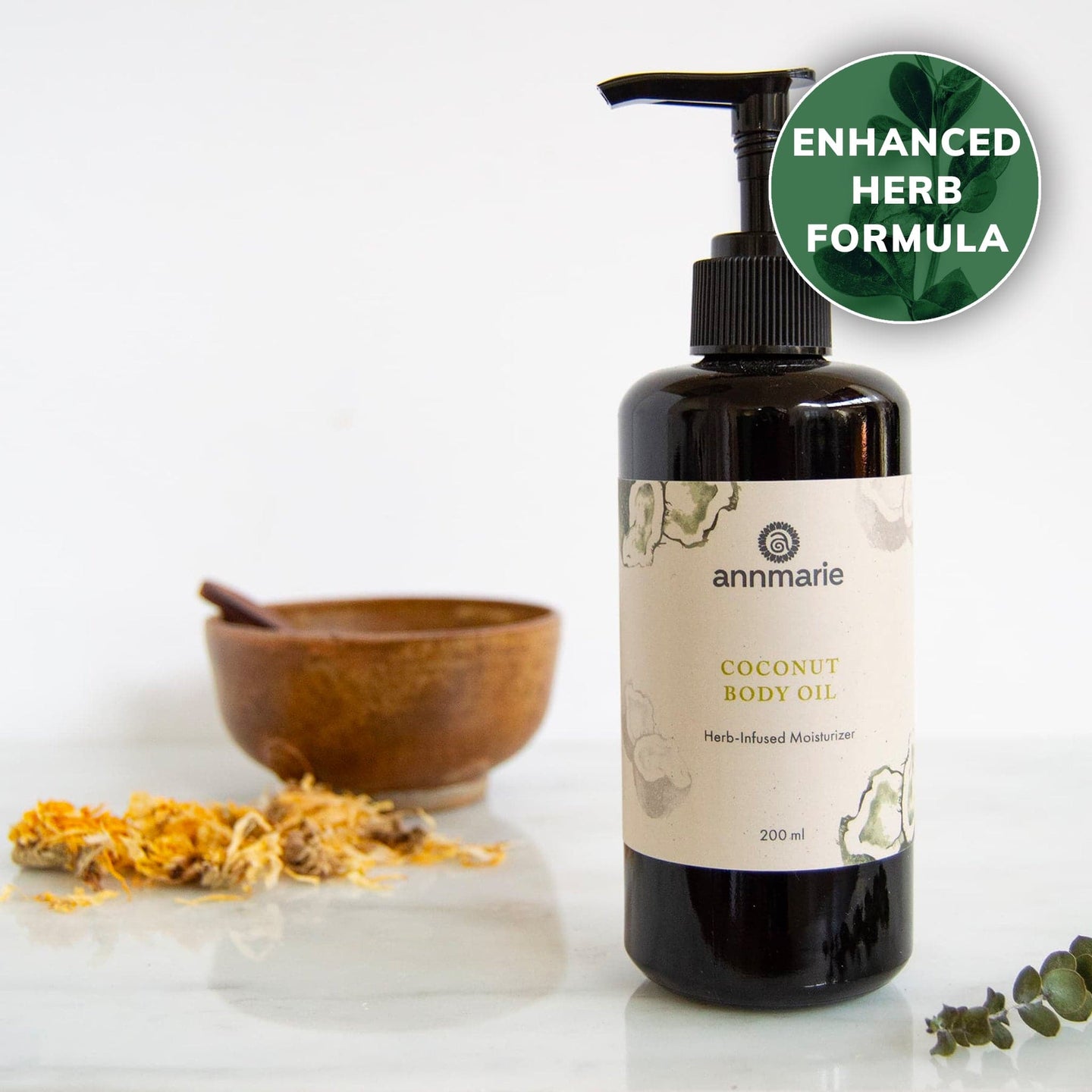 Annmarie Coconut Body Oil, an herb-infused moisturizer, sits beside calendula petals and a wooden bowl on a white marble surface.
