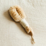 A natural bristle dry brush with a jute-wrapped handle sits on a textured linen surface.
