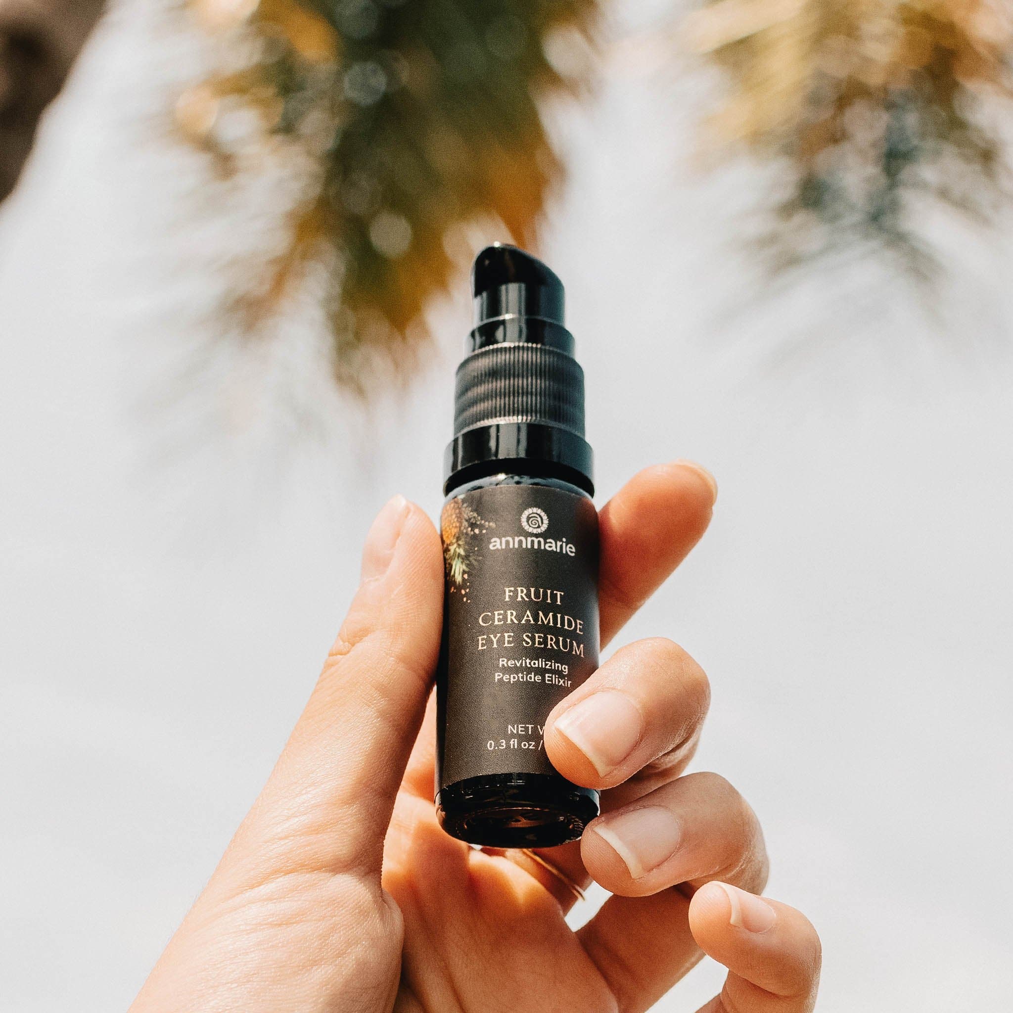 A hand holds Annmarie Fruit Ceramide Eye Serum against a bright, outdoor background.
