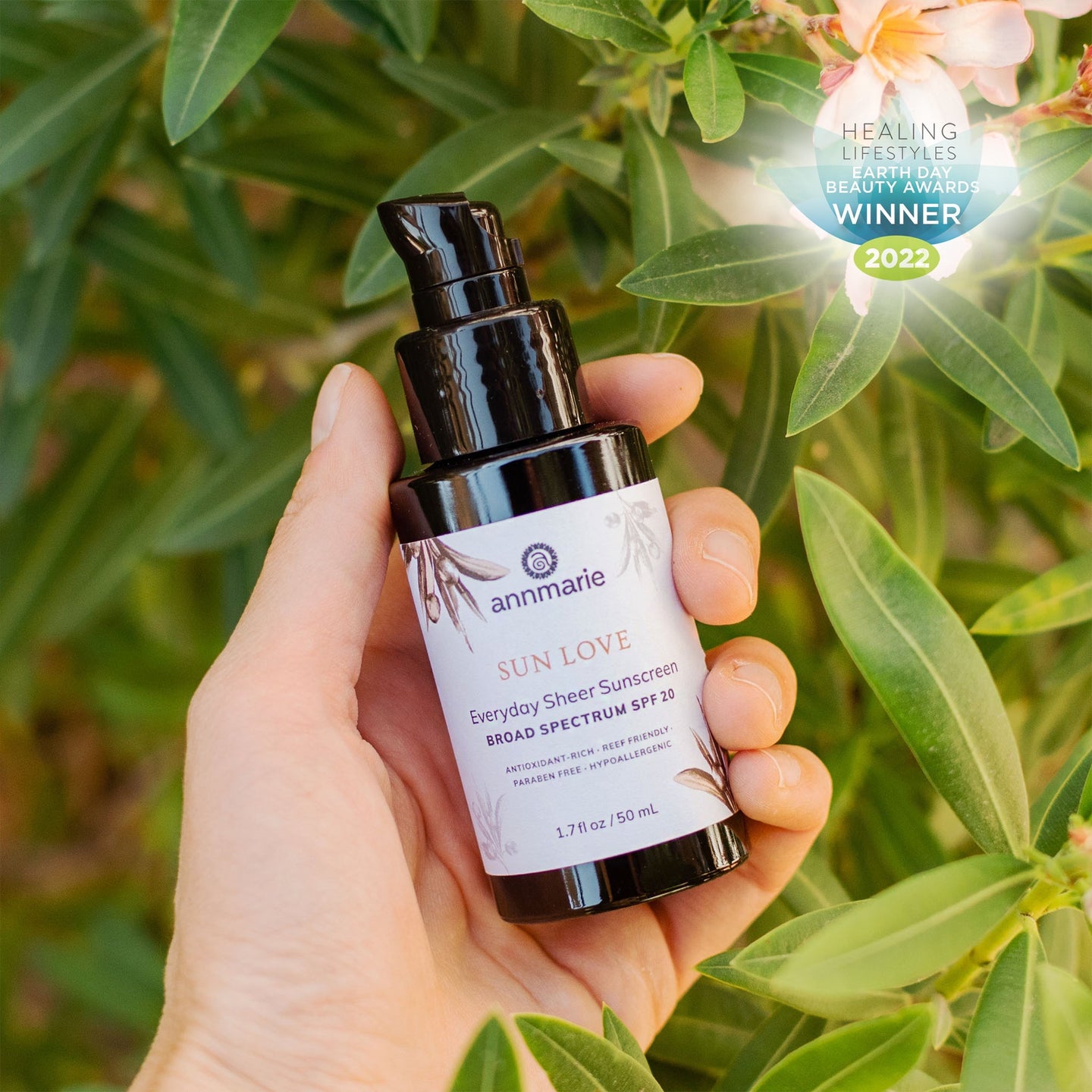 A hand holds Annmarie Sun Love everyday sheer sunscreen SPF 20, an award-winning reef-friendly beauty product, against a backdrop of green leaves and flowers.
