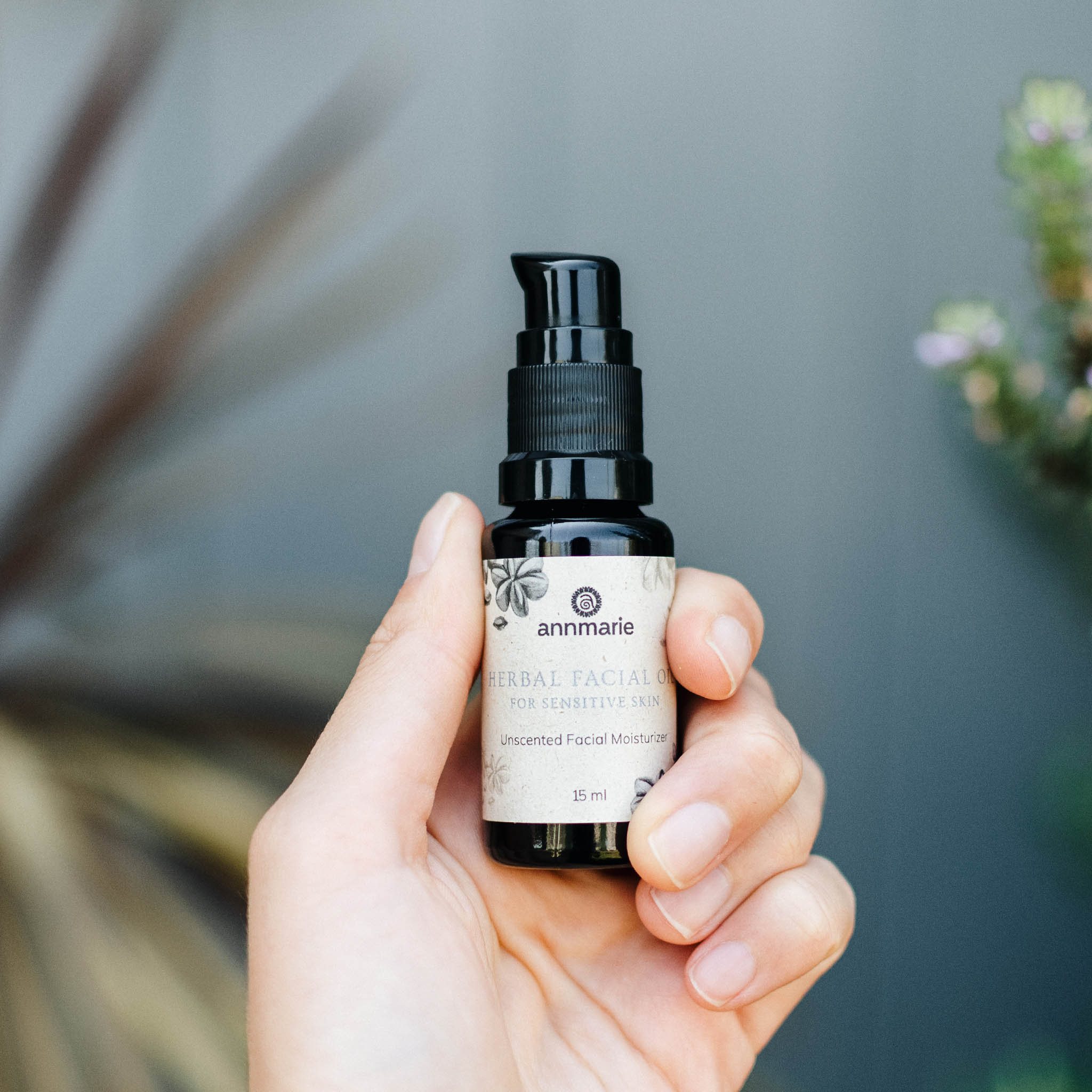 A hand holds a bottle of Annmarie Herbal Facial Oil, an unscented moisturizer for sensitive skin.
