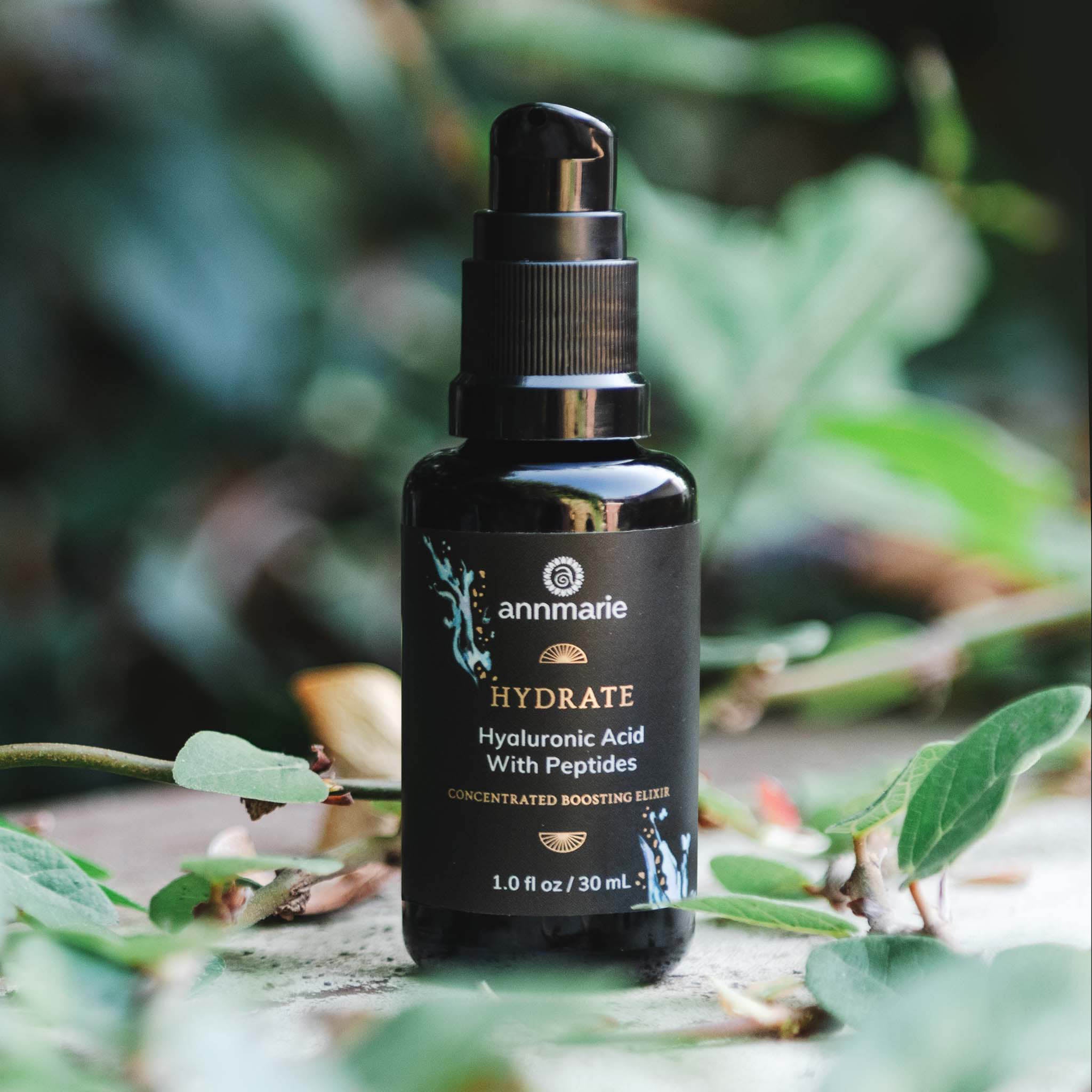 Annmarie's HYDRATE skincare serum, a hyaluronic acid and peptide boosting elixir, sits outdoors amongst green foliage.
