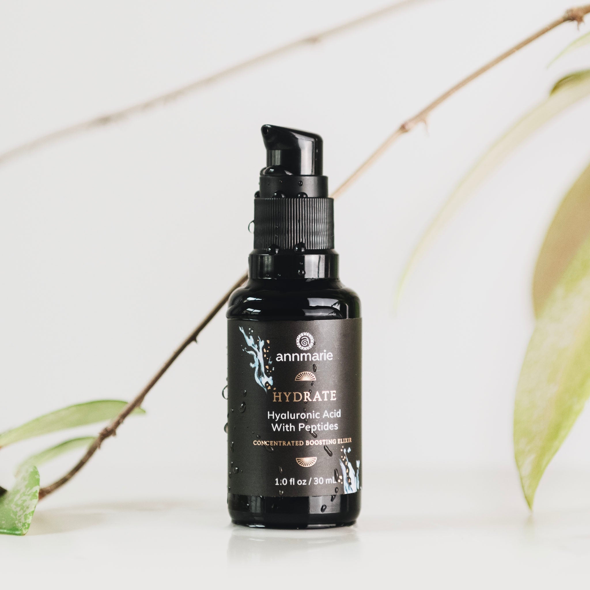 Annmarie's black Hyaluronic Acid and Peptides elixir bottle promotes skin hydration against a botanical backdrop.
