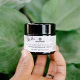 A hand holds a jar of Annmarie Illuminating Pearl Mask, a hydrating and brightening skin treatment, against a lush green background.
