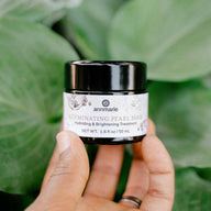 A hand holds a jar of Annmarie Illuminating Pearl Mask, a hydrating and brightening skin treatment, against a lush green background.

