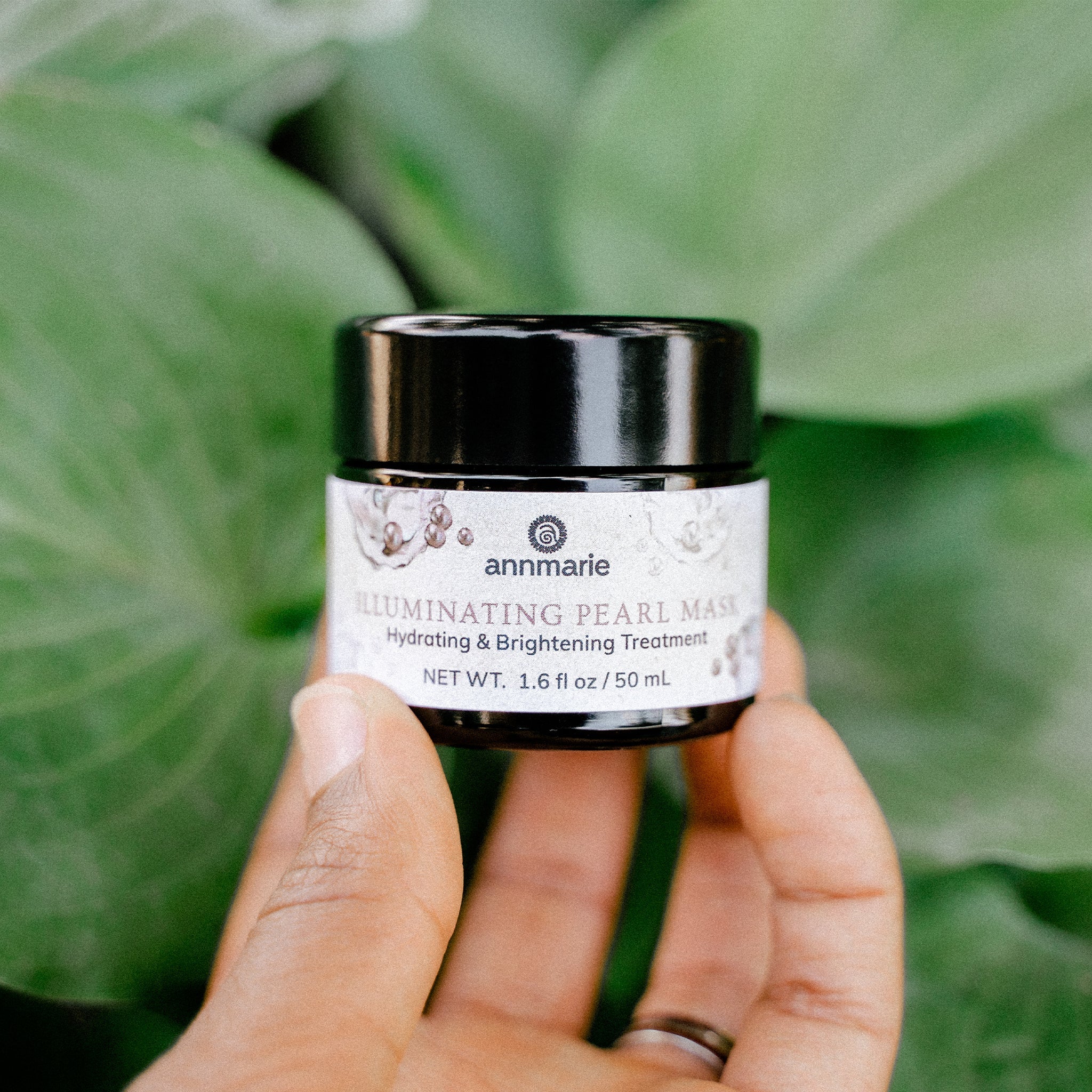 A hand holds a jar of Annmarie Illuminating Pearl Mask, a hydrating and brightening skin treatment, against a lush green background.
