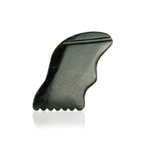 A jade gua sha tool with a toothed edge rests on a reflective white surface.
