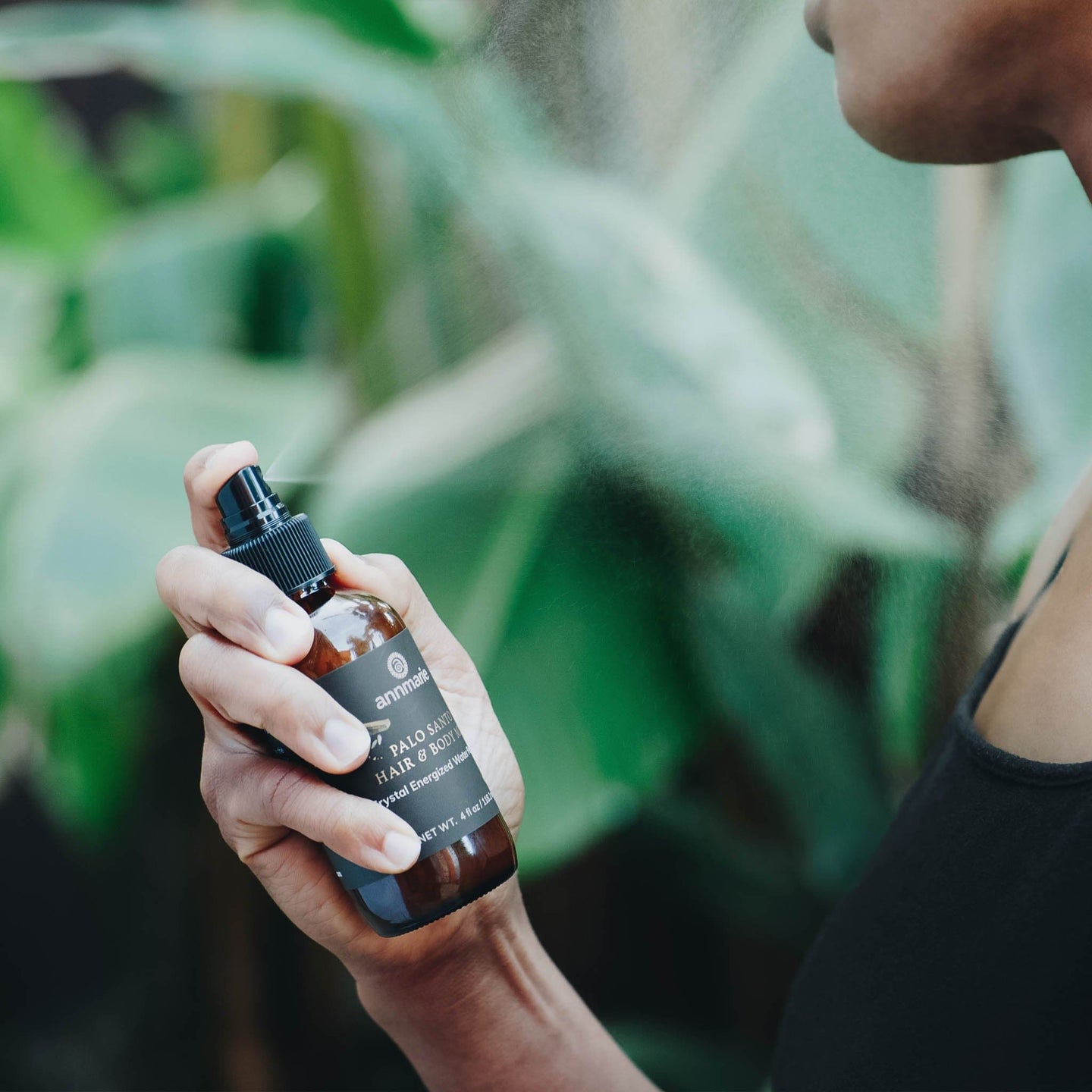 A person sprays Annmarie Palo Santo hair and body mist against a green, leafy background.
