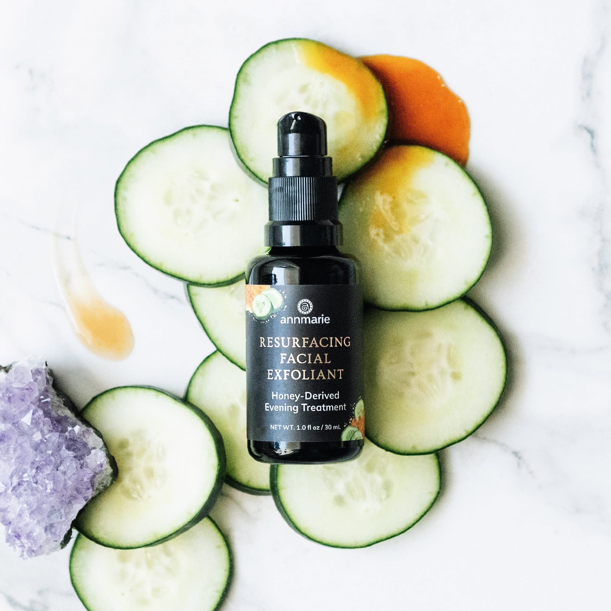 Annmarie's Resurfacing Facial Exfoliant, a honey-derived evening treatment, sits among fresh cucumber slices and an amethyst crystal on a marble surface.
