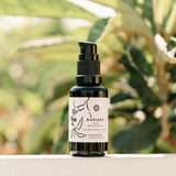 Radiate Facial Moisturizing Oil sits on a ledge against a blurred leafy background, showcasing its natural beauty product label.
