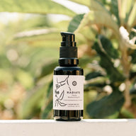 Radiate Facial Moisturizing Oil sits on a ledge against a blurred leafy background, showcasing its natural beauty product label.
