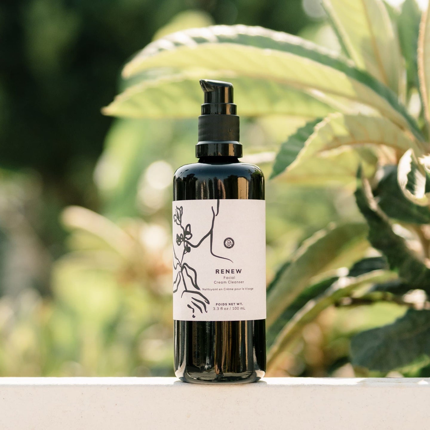 A black bottle of RENEW facial cream cleanser sits on a wall with lush green foliage in the background.
