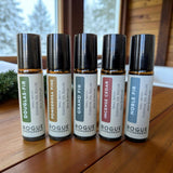 Five Rogue Evergreens essential oil rollers, featuring Douglas Fir, Ponderosa Pine, Grand Fir, Incense Cedar, and Noble Fir, stand on a wooden table.
