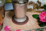 A frosted silver mint julep cup rests on a leather coaster alongside a pink rose and a horse racing trophy.
