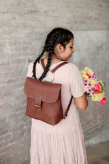 Woman with braids wears a leather backpack and carries a colorful bouquet of flowers.

