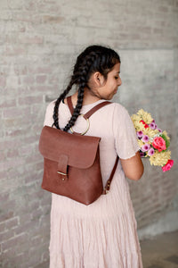 Woman with braids wears a leather backpack and carries a colorful bouquet of flowers.
