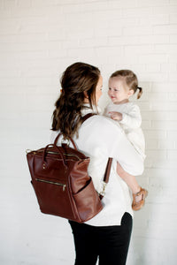 A mother, wearing a stylish brown leather diaper bag backpack, smiles lovingly at her young daughter whom she's holding.

