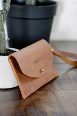 Close-up features an Urban Southern leather wristlet wallet resting on a wooden surface near a potted plant.
