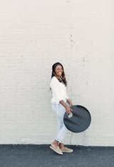 Smiling woman holds a stylish, round, black leather handbag against a textured brick wall.
