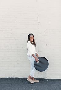 Smiling woman holds a stylish, round, black leather handbag against a textured brick wall.
