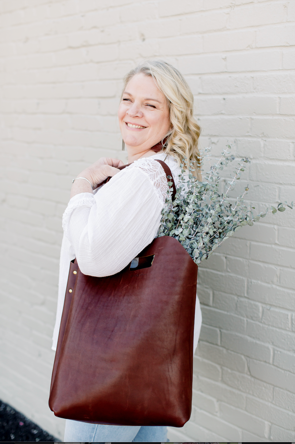 Smiling woman carries a stylish brown leather tote bag filled with eucalyptus against a white brick wall.

