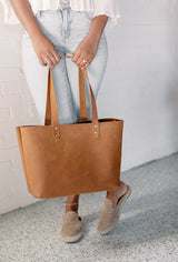A woman showcases a stylish brown leather tote bag with riveted straps, paired with light-wash jeans and animal-print mules.
