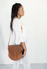 Woman models a stylish brown leather saddle bag with braided hair, white blouse, and light wash jeans.
