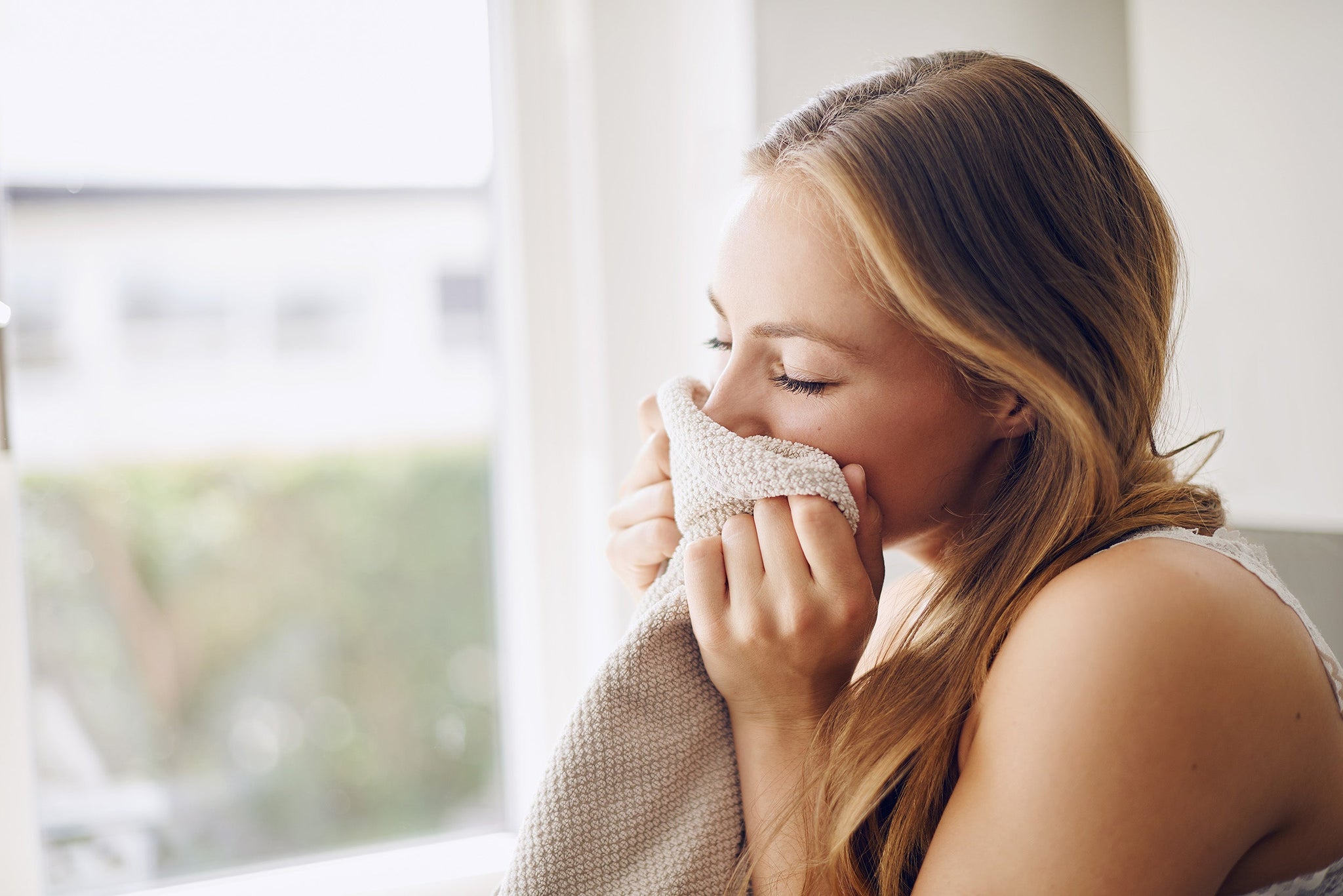 A young woman with blonde hair gently presses a soft, beige towel to her face while standing by a bright window.
