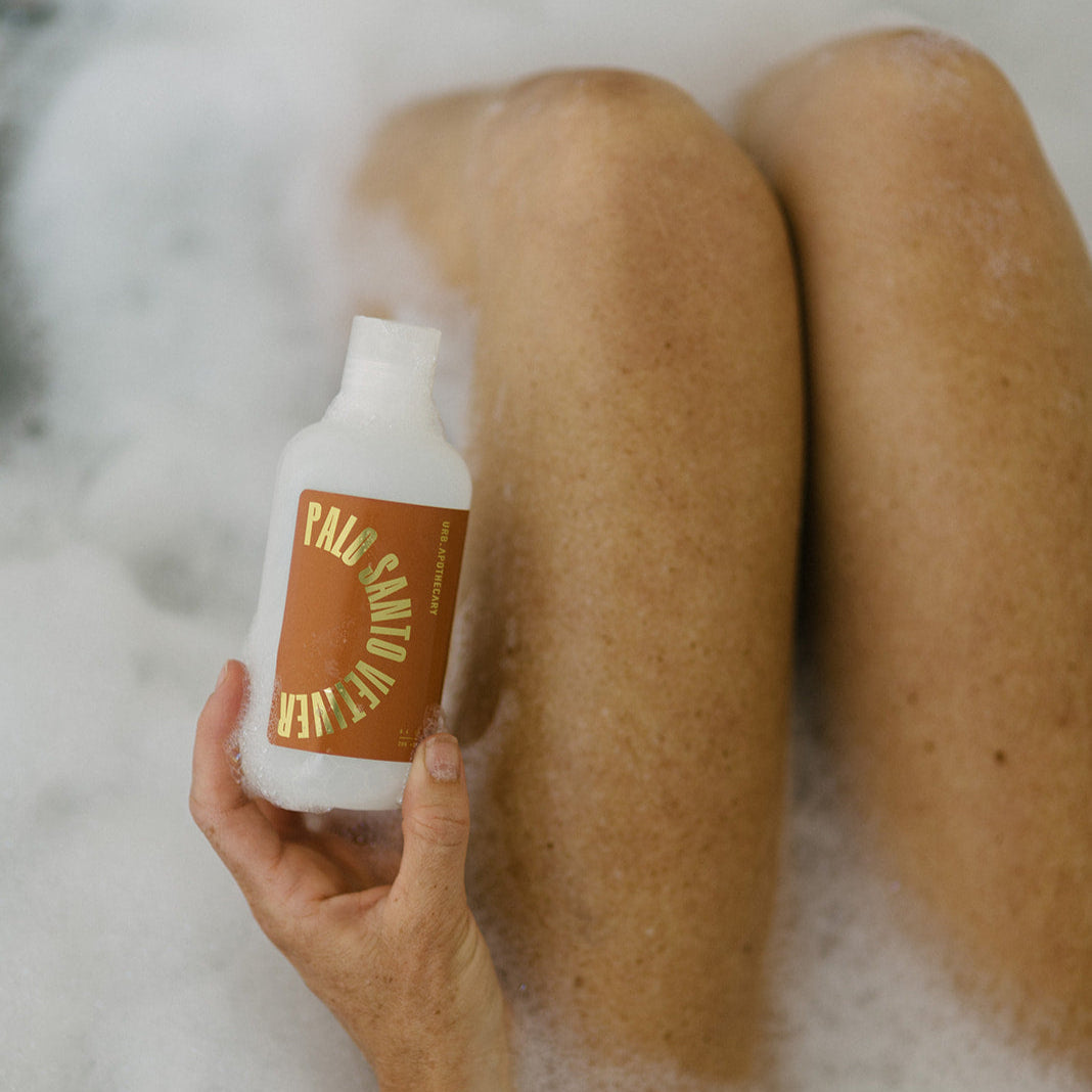 A woman soaks in a bubble bath while holding a bottle of Palo Santo Vetiver bath soak.
