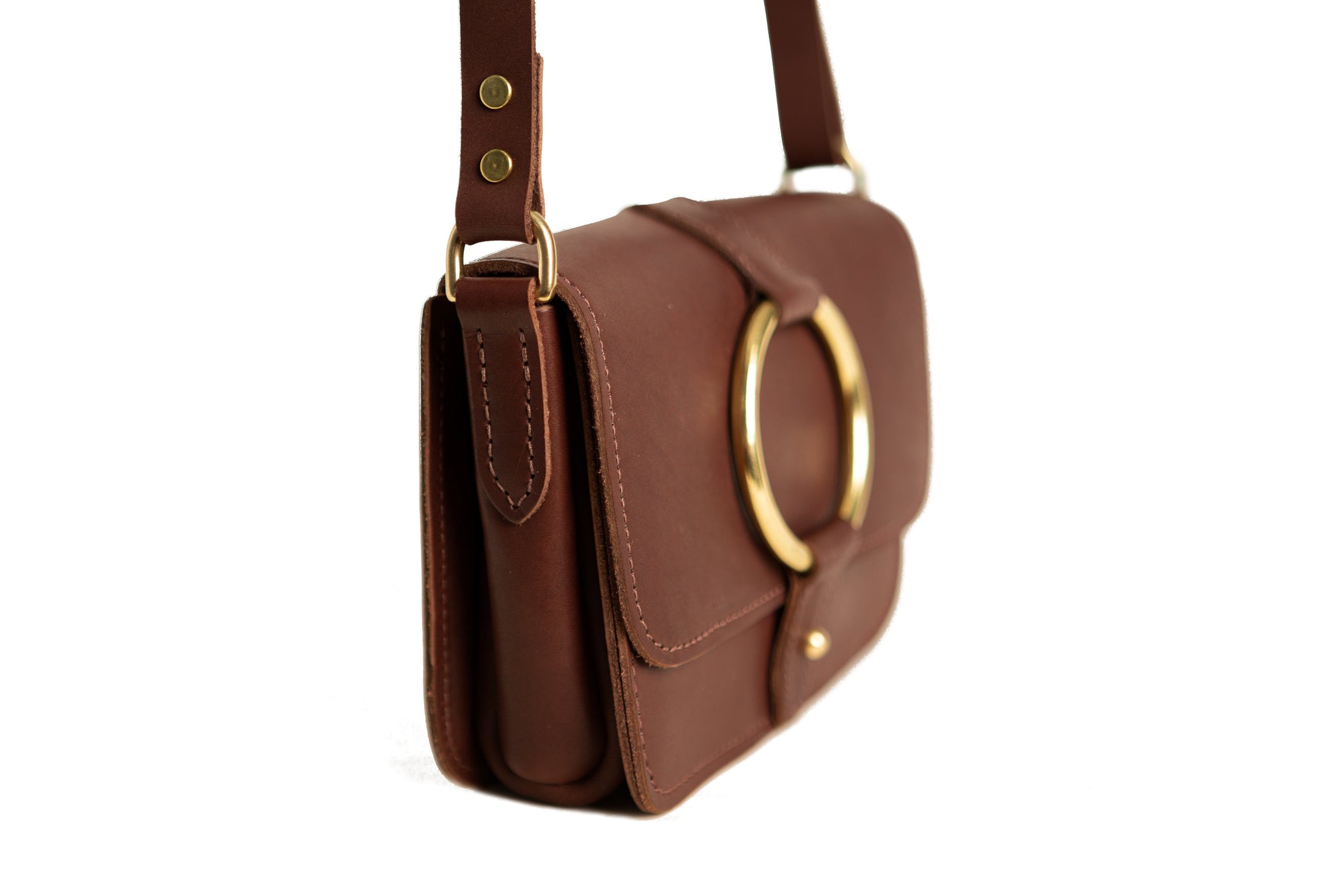 A brown leather saddlebag purse with a gold ring clasp hangs against a white background.
