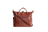 Displaying a brown leather tote bag with top handles and a crossbody strap against a white background.
