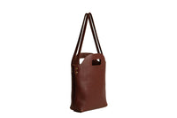 A stylish brown leather tote bag with long handles stands isolated on a white background.
