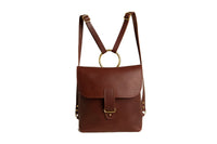 Showcases a stylish brown leather backpack purse featuring gold hardware and a circular handle against a clean white background.
