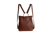 A brown leather backpack features a gold circular handle and adjustable straps, presented against a bright white background.
